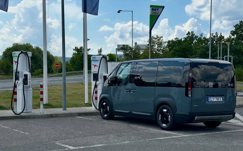 The grey van plugged in at a fast-charging station on a sunny day.