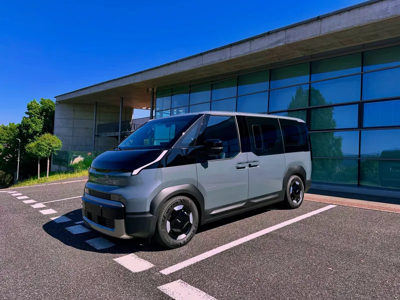 The grey van with a black bonnet parked in an empty car park in front of a glass-fronted building.