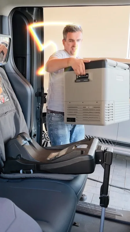 A man lifts a grey portable fridge into the van through the side door, past a child-seat base on the rear seat.