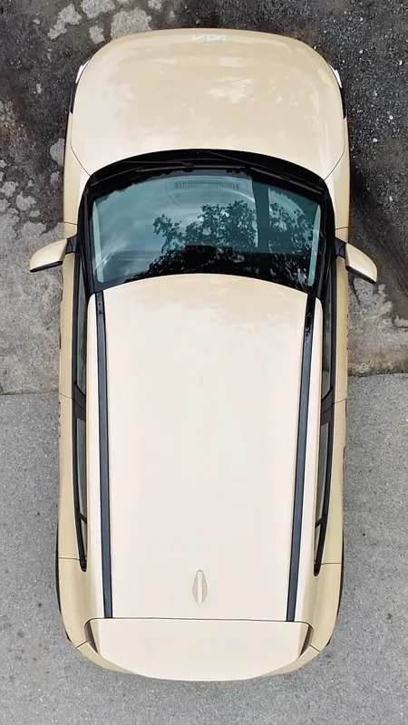 Top-down drone view of a small beige car with black roof rails parked on grey asphalt.