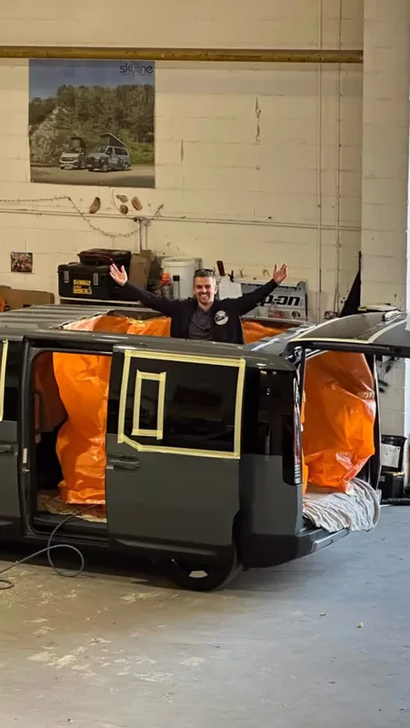 In a workshop, a man stands with raised arms in a large hole cut in the van’s roof, the interior covered in orange sheeting.