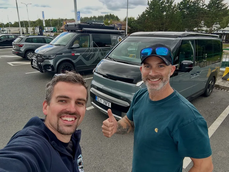 Two smiling men take a selfie in a car park in front of the grey camper van and a dark camper with a roof rack.