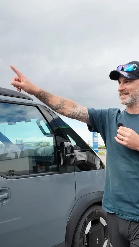A bearded man in a cap and sunglasses points across the grey van in a car park under a cloudy sky.