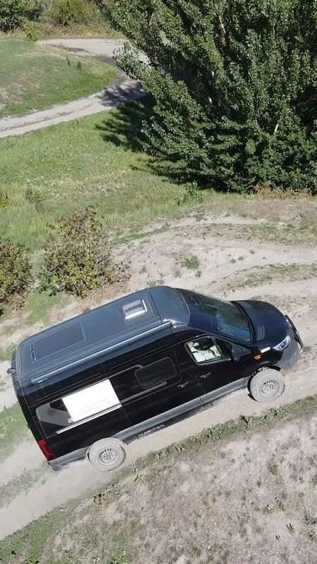 Drone view of a black camper van driving down a dusty off-road track past green bushes.