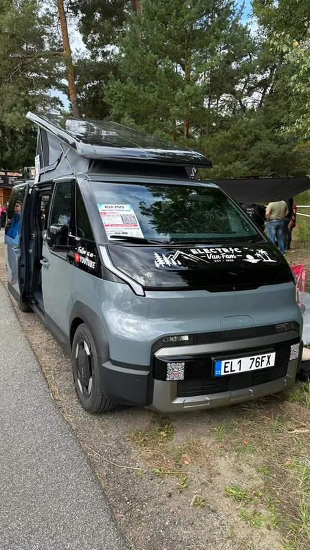 The grey camper van with its pop-top roof raised and side door open, parked among pine trees at a meet-up.