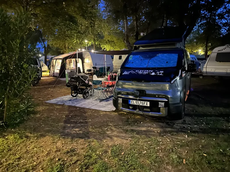 The van parked at a campsite in the evening, with an awning, chairs and a pram