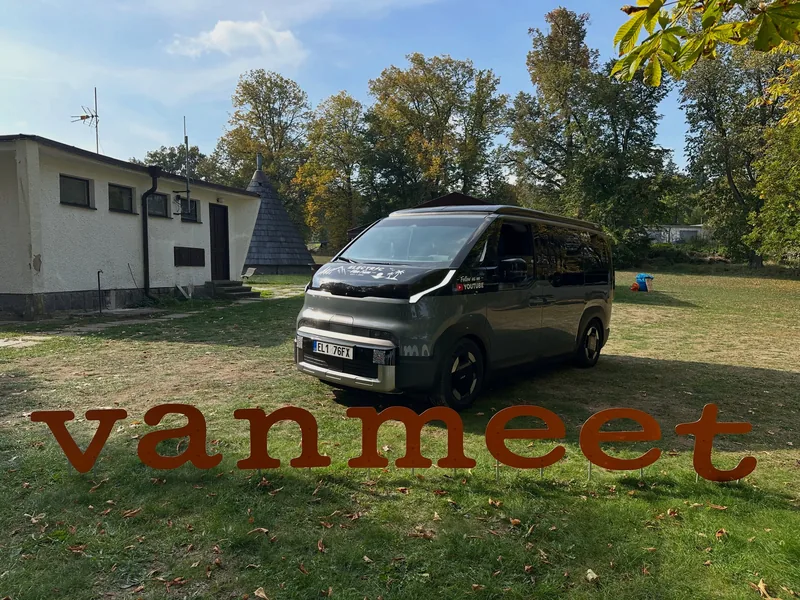 The grey camper van parked on a sunny lawn behind large rust-coloured letters spelling “vanmeet”.